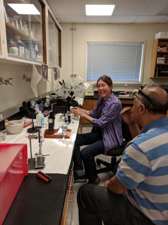 Two people in a lab, one smiling at a microscope, another listening attentively.